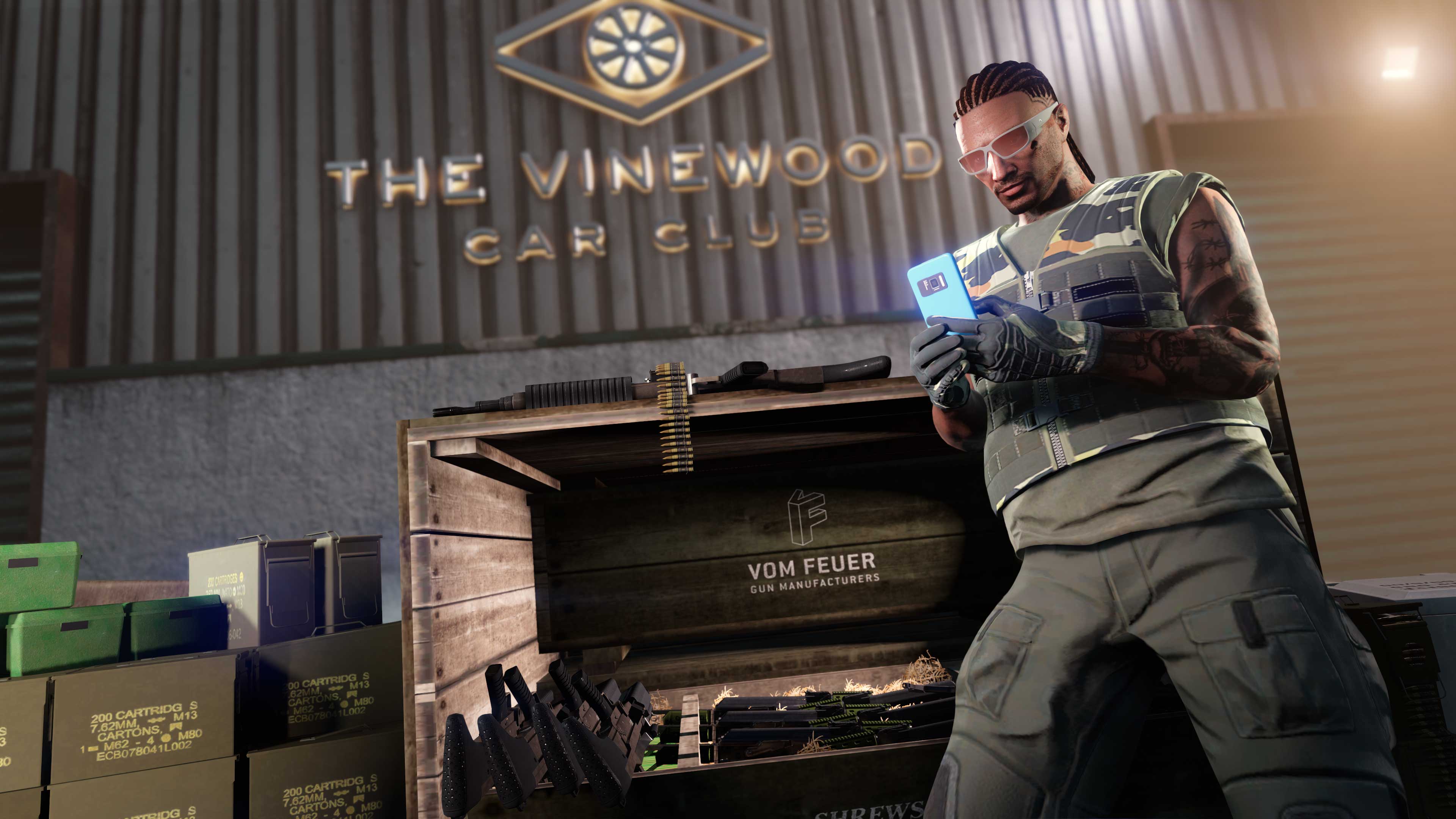 A GTA Online character on his phone leans against a crate full of weapons in front of The Vinewood Car Club. 